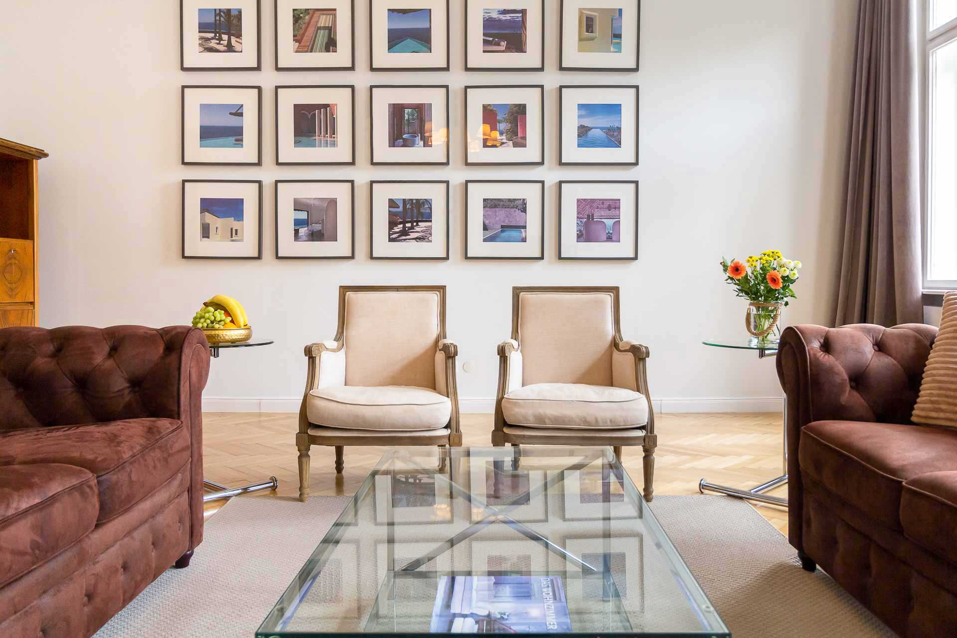 2 charming and historic chairs in the center , on the left and right side 2 elegant brown couches, in the middle a minimalistic glass table, colorful pictures in the wallin the back; room is located in the 8th district in Vienna
